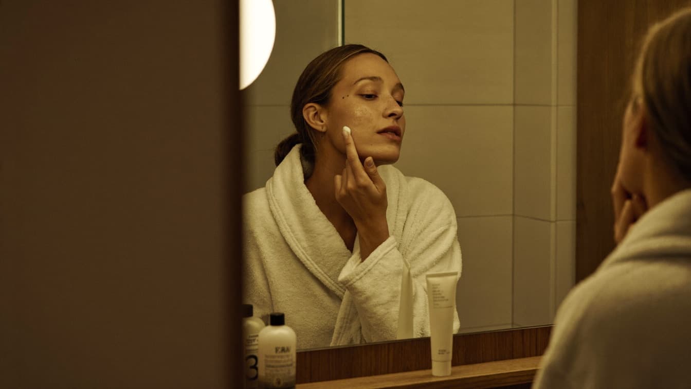 A person applying a pea-sized amount of prescription cream to clean dry skin in front of a bathroom mirror at night